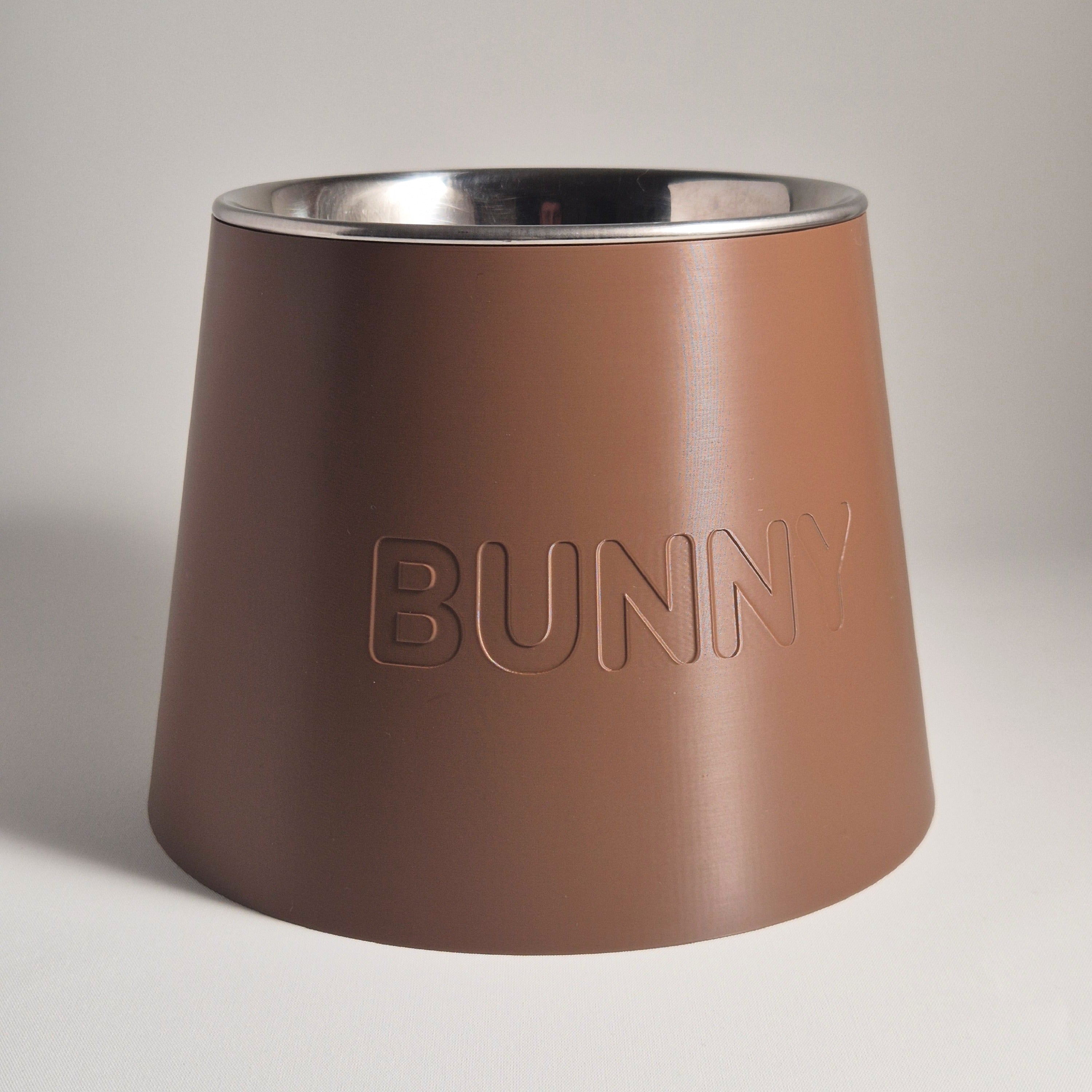 A conical pet feeder or bowl with a matte brown exterior and a polished stainless steel interior, featuring the word 'BUNNY' embossed on the front, set against a neutral light gray background.
