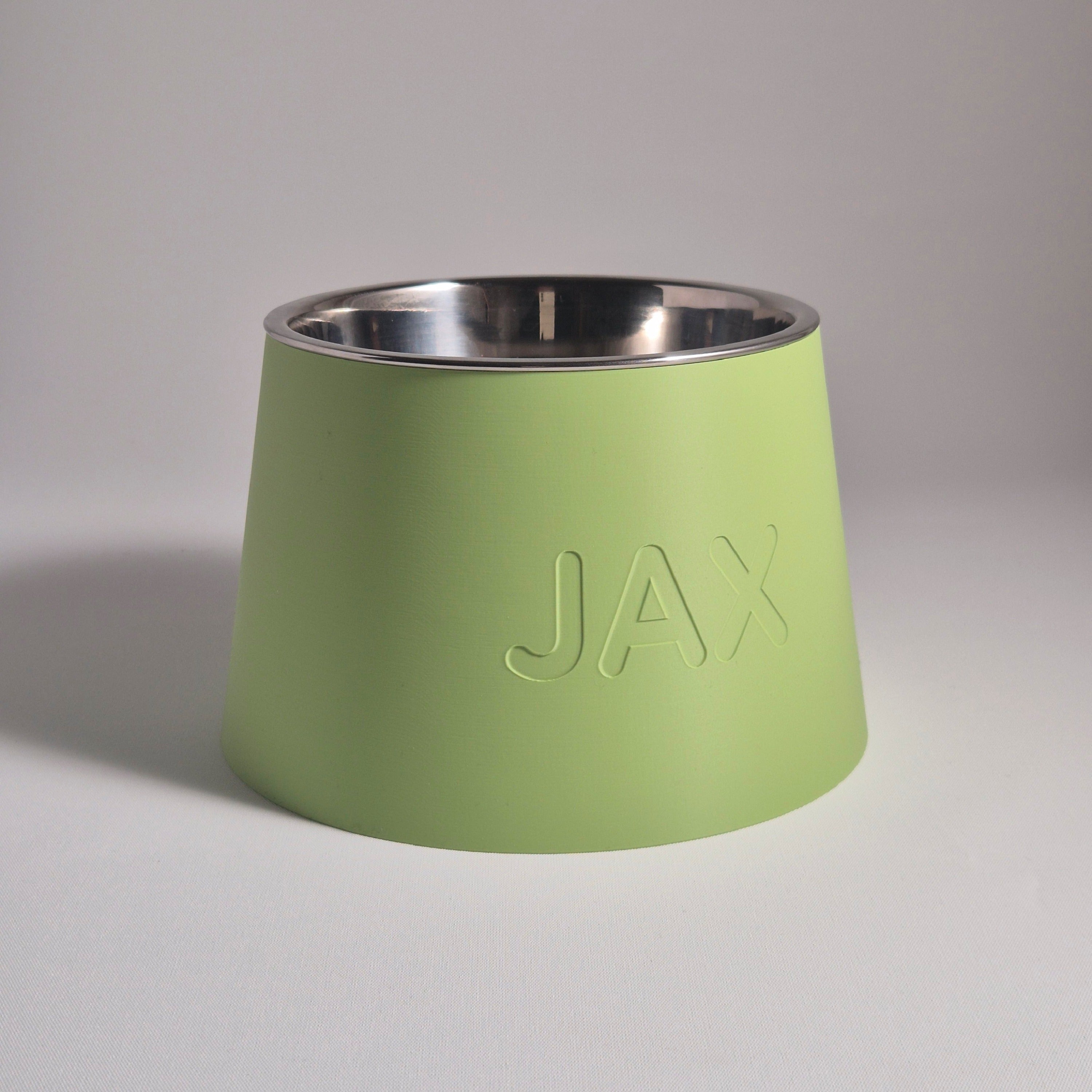 A light green pet bowl with a stainless steel interior and the name 'JAX' embossed on the front, set against a neutral gray background.