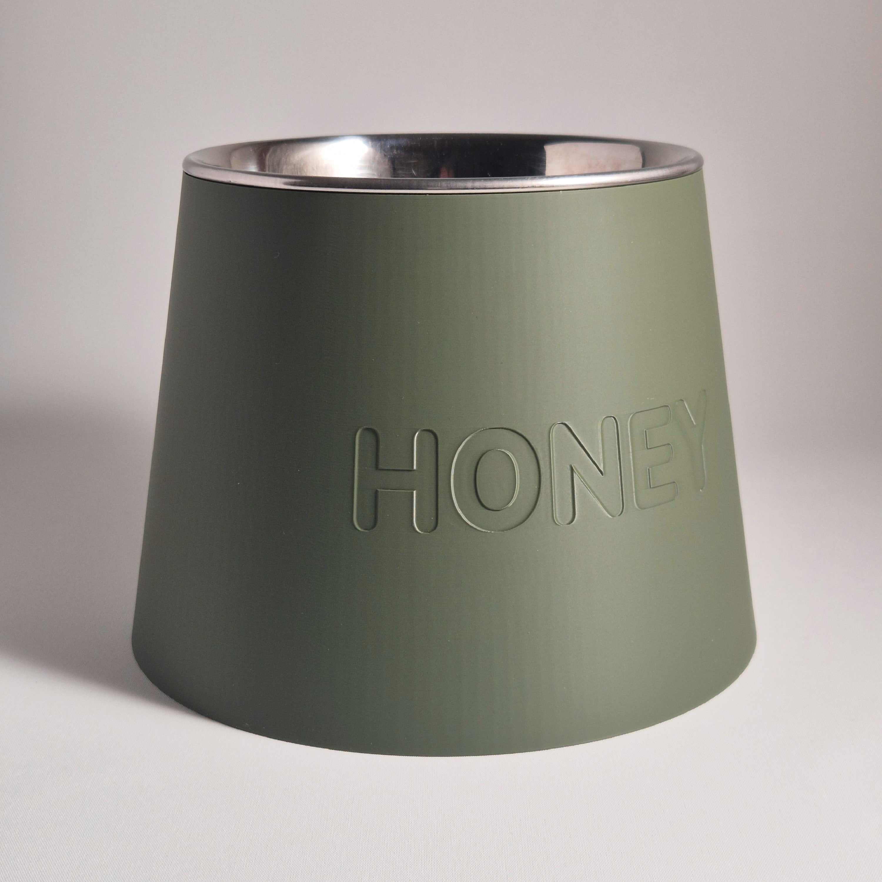 An olive green pet feeder bowl with a brushed silver stainless steel interior, featuring the word 'HONEY' embossed on the front, set against a neutral light gray background.