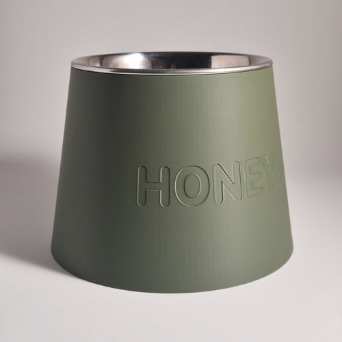 An olive green pet feeder bowl with a brushed silver stainless steel interior, featuring the word 'HONEY' embossed on the front, set against a neutral light gray background.