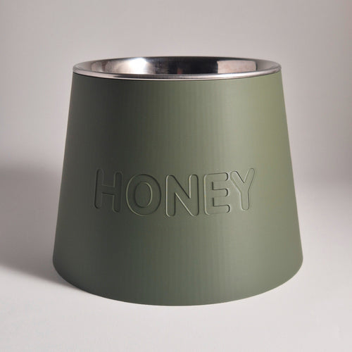An olive green conical pet bowl with a brushed silver stainless steel interior, featuring the word 'HONEY' embossed on the front, set against a neutral gray background.