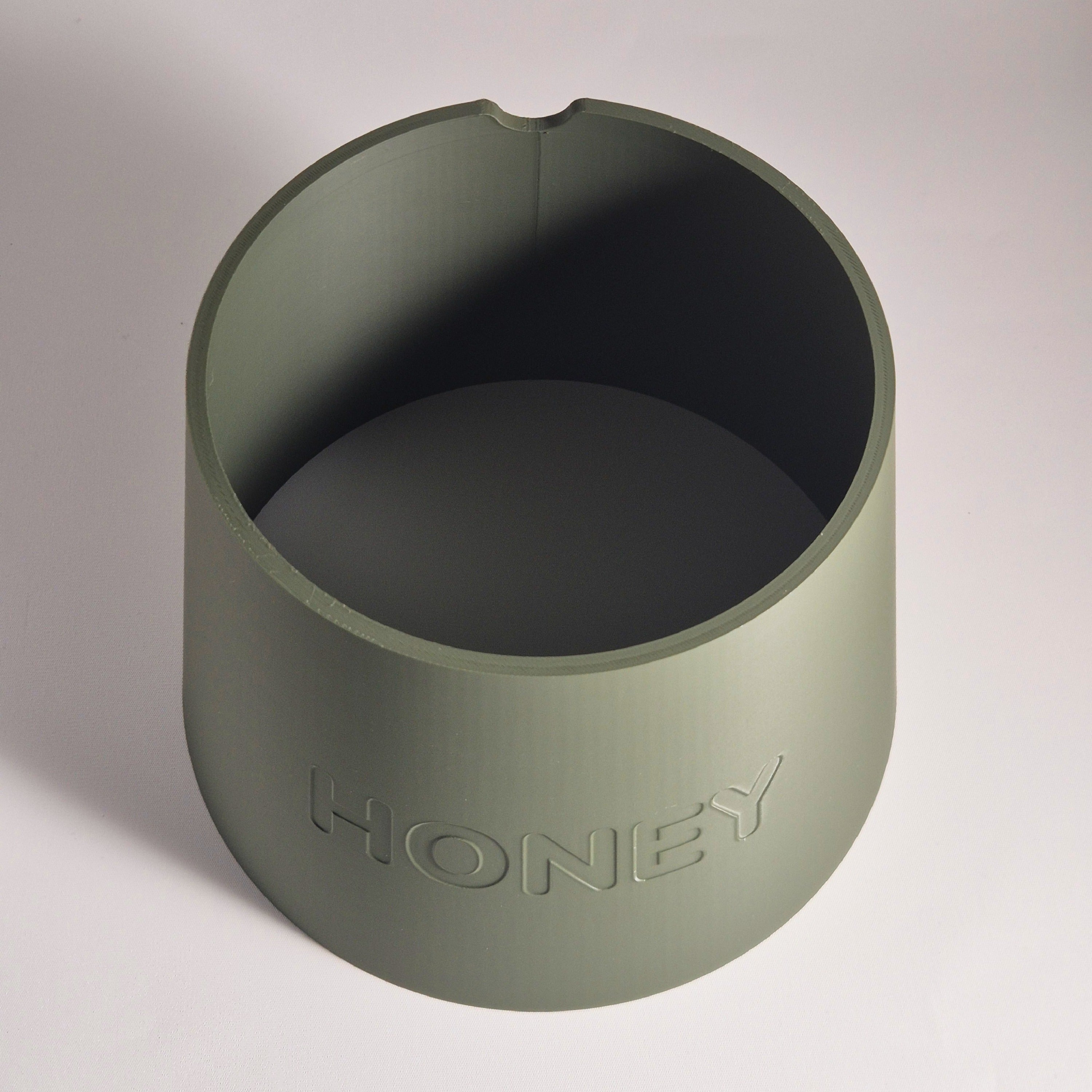 An olive green cylindrical container with a small notch at the top rim, featuring the word 'HONEY' embossed on the front, set against a neutral light gray background.